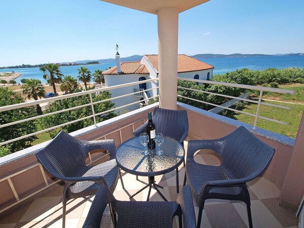 Un balcon avec des fauteuils en osier et une table, offrant une vue sur la côte, avec un bâtiment et des palmiers.