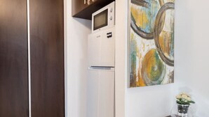 Eine kompakte Küche mit einem weißen Kühlschrank, einer Mikrowelle und einem kleinen Schrank darüber. An der Wand hängt ein Bild und auf einem Holznachttisch steht eine Vase mit Blumen.