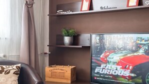 Ein modernes Wohnzimmer mit einem Flachbildfernseher, der einen Filmplakat zeigt, einem Glastisch mit Dekoration, einem Ledersofa und einer an der Wand befestigten Regalanlage mit Dekorationselementen.