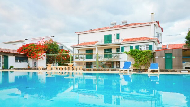A swimming pool with clear blue water, surrounded by a building with a red-tiled roof and green shutters.