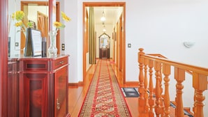 A hallway with a wooden staircase, a red carpet, and a mirror reflecting a door and a room with a chandelier.