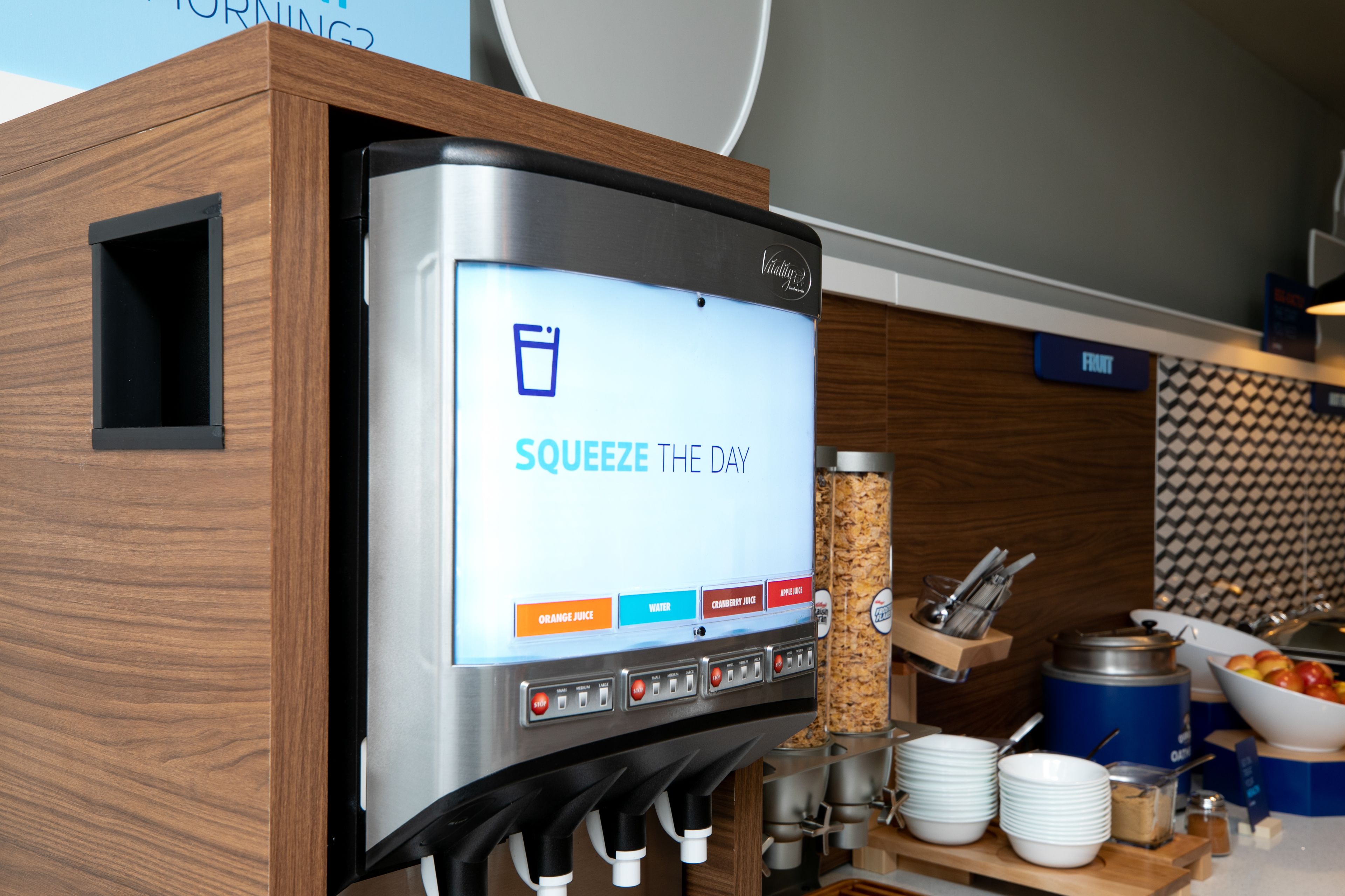 A beverage dispenser with a digital screen displaying options for orange juice, water, cranberry juice, and apple juice.