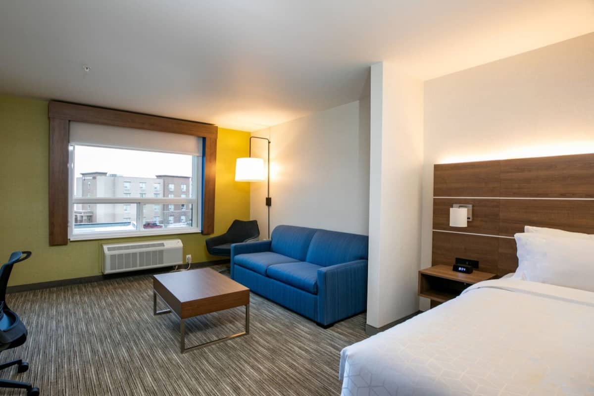 A hotel room with a bed, a blue sofa, a small table, a desk with a chair, a window with a view of buildings, and a wall-mounted lamp.