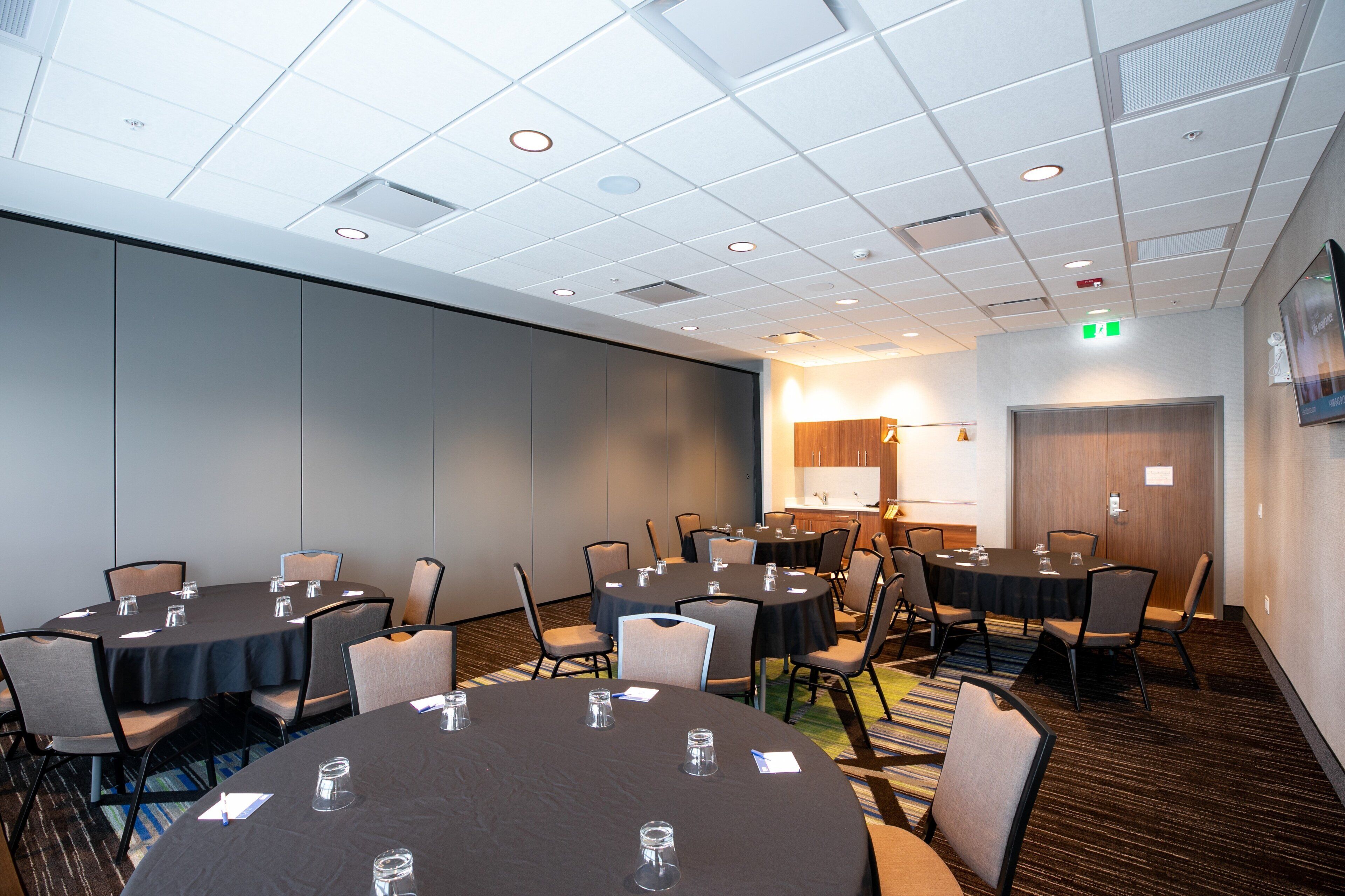 A conference room with multiple tables and chairs, a kitchenette, and a door with an emergency exit sign.