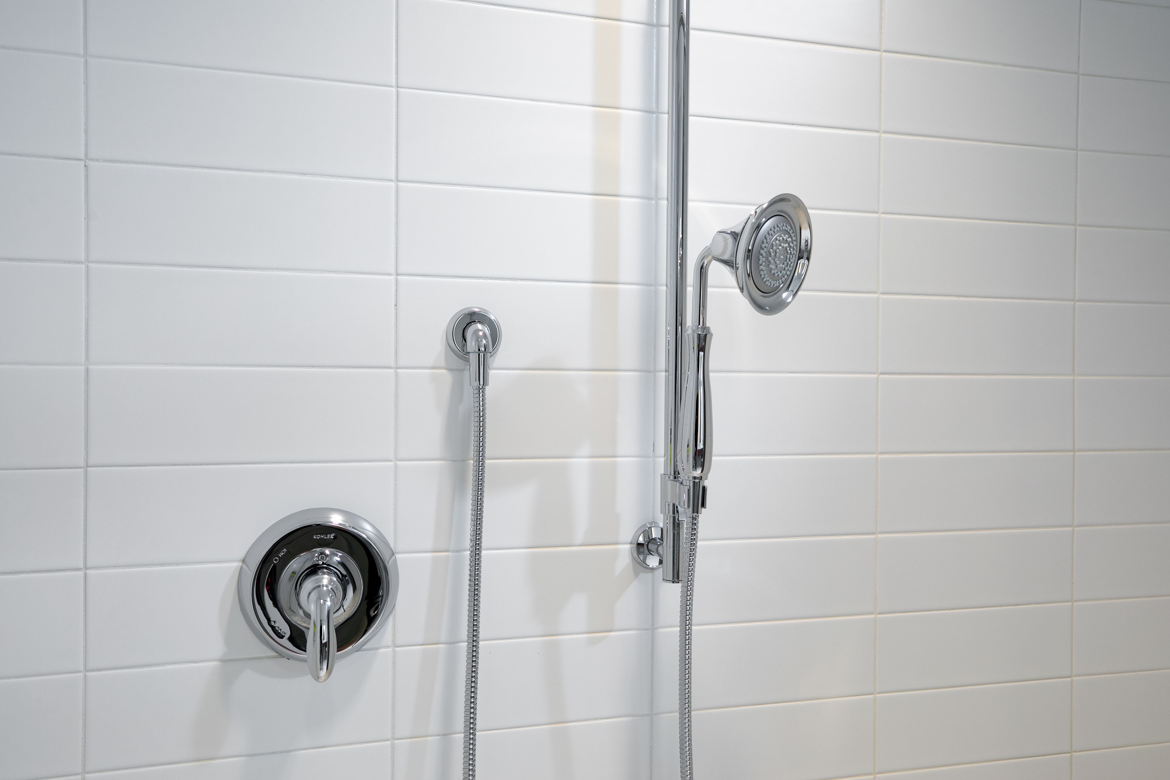 Un baño con paredes de azulejos blancos, una ducha con cabezal de ducha manual y un grifo empotrado.
