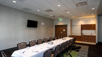 A conference room with a long table, chairs, a flat-screen TV, and a kitchenette area.