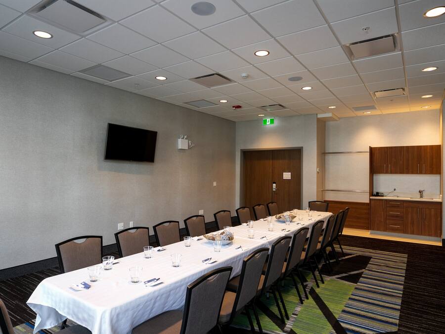 Un salón de conferencias con una larga mesa, sillas, un televisor de pantalla plana y una zona de cocina.