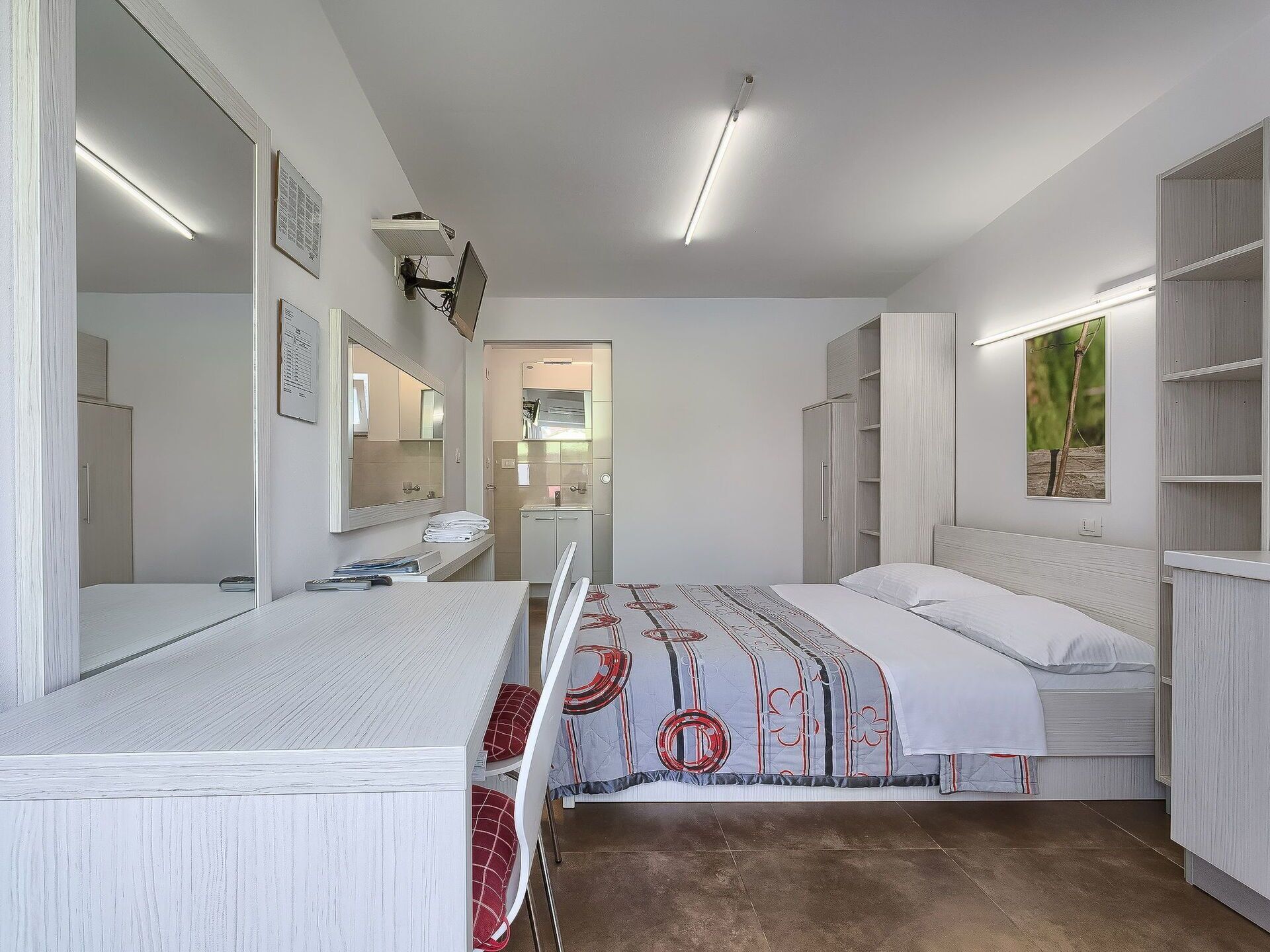 Ein modernes Hotelzimmer mit einem großen Bett, einem Schreibtisch, einem Spiegel und einem Kleiderschrank.