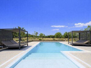 A rectangular swimming pool with two covered lounging areas on either side, surrounded by a landscaped garden with trees and a clear blue sky.