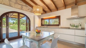 Una cocina con pared de piedra, vigas de madera, lámpara colgante, ventana con vistas y una mesa con sillas.