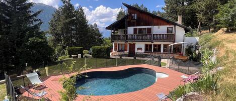 A two-story chalet with a swimming pool, surrounded by trees and mountains.