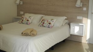 Una cama bien hecha con almohadillas florales, una manta blanca y toallas enrolladas. Hay dos mesitas de noche con lámparas y una luz empotrada en la pared.