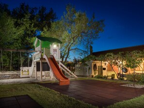 Um parquinho com um escorrega e uma estrutura de escalada, rodeado por árvores e um edifício com telhado de cerâmica.