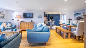 A modern living room with a sofa, a coffee table, and a dining area with a bench and chairs. There is a kitchen in the background with cabinets and a television mounted on the wall. - Stone Lodge, Old Hunstanton, Norfolk (Old Hunstanton)