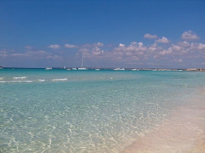 A beach with clear turquoise water, a sandy shore, and a distant sailboat.