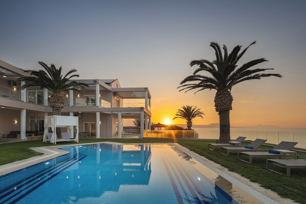 A pool area with a sunset view, lounge chairs, and a building with a covered seating area.