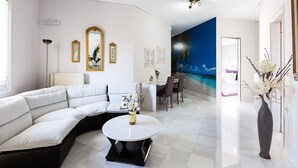 Een moderne woonkamer met een witte modulaire bank, een ronde salontafel en een eethoek met tafel en stoelen. Op de muur hangt een groot schilderij van een strand, en op een bijzettafeltje staat een vaas met bloemen.