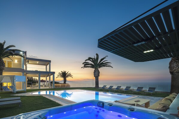 A modern pool area with lounge chairs and a hot tub, overlooking a body of water and a sunset.