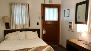Ein Hotelzimmer mit Bett, Schreibtisch, Lampe, Tür und einem Fenster mit Vorhängen.