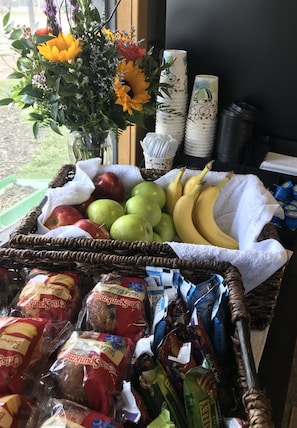 A table with a basket of apples, bananas, and bread, a vase with flowers, and stacks of paper cups and plates.