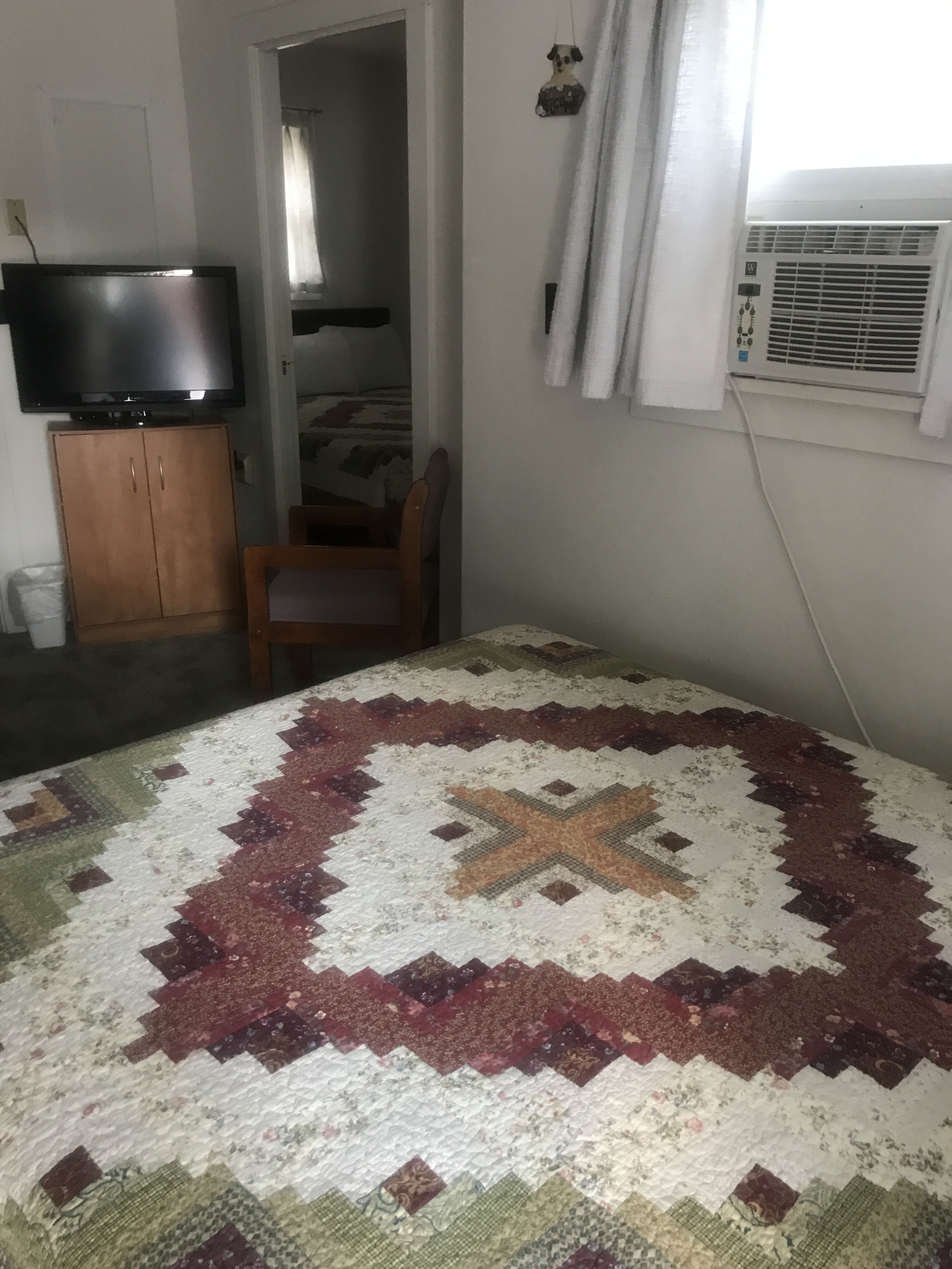 Una habitación con edredón acolchado, televisor sobre un mueble de madera, una silla y un equipo de aire acondicionado.