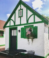 A small white building with green trim and shutters, a black door, and a small table with chairs in front.