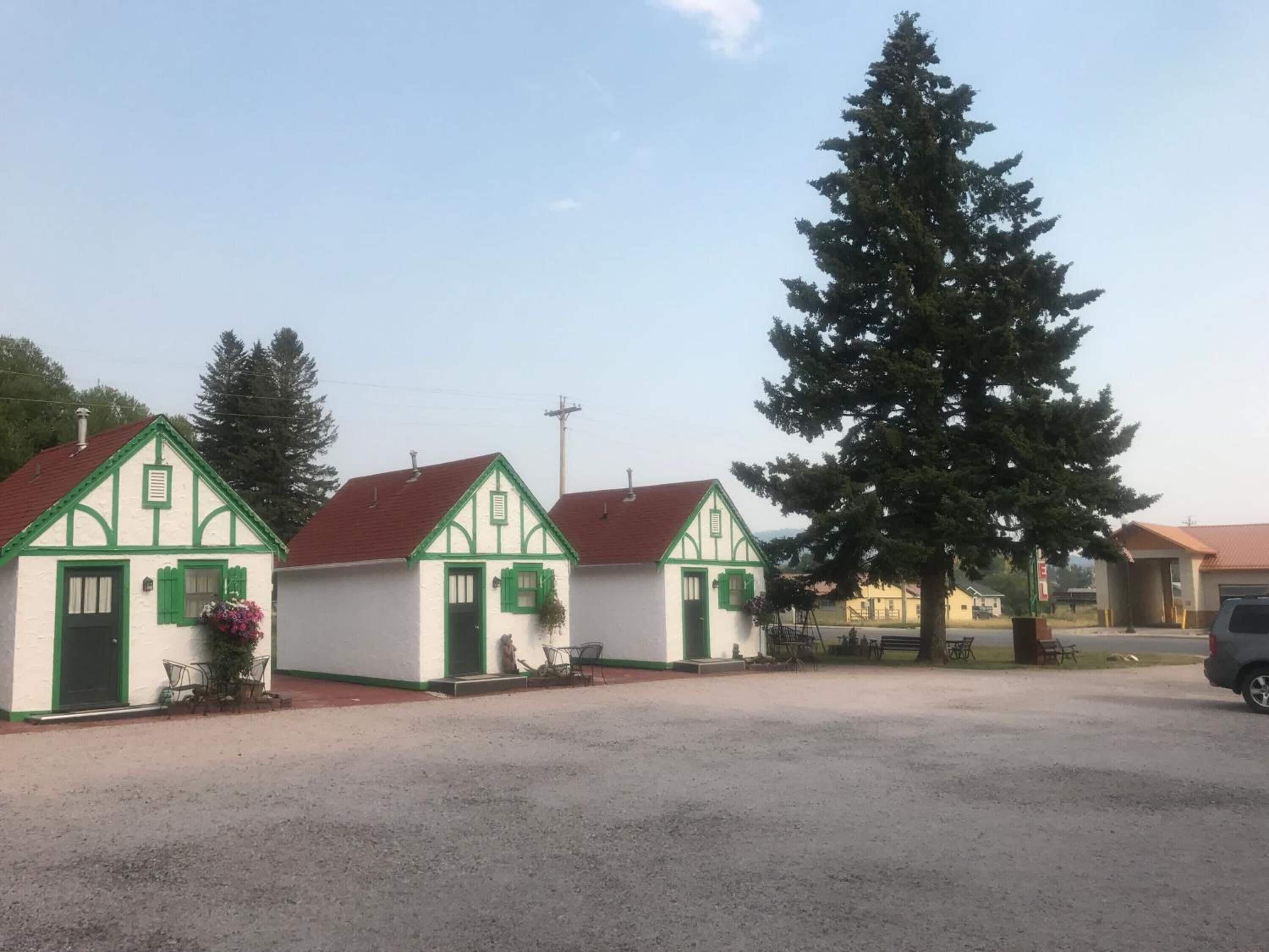 Une rangée de petits bâtiments blancs, agrémentés de bordures vertes et surmontés de toits rouges, qui sont probablement des maisons d’hôtes ou des chalets, avec un grand arbre à feuilles persistantes devant.