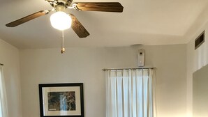 Ein Zimmer mit einem roten Sofa, einem Holztisch, einer Lampe, einem Sessel, einem Deckenventilator, einem gerahmten Bild und einem Fenster mit Vorhängen.