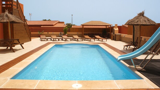 A swimming pool with lounge chairs and a slide, surrounded by wooden structures and a clear sky.