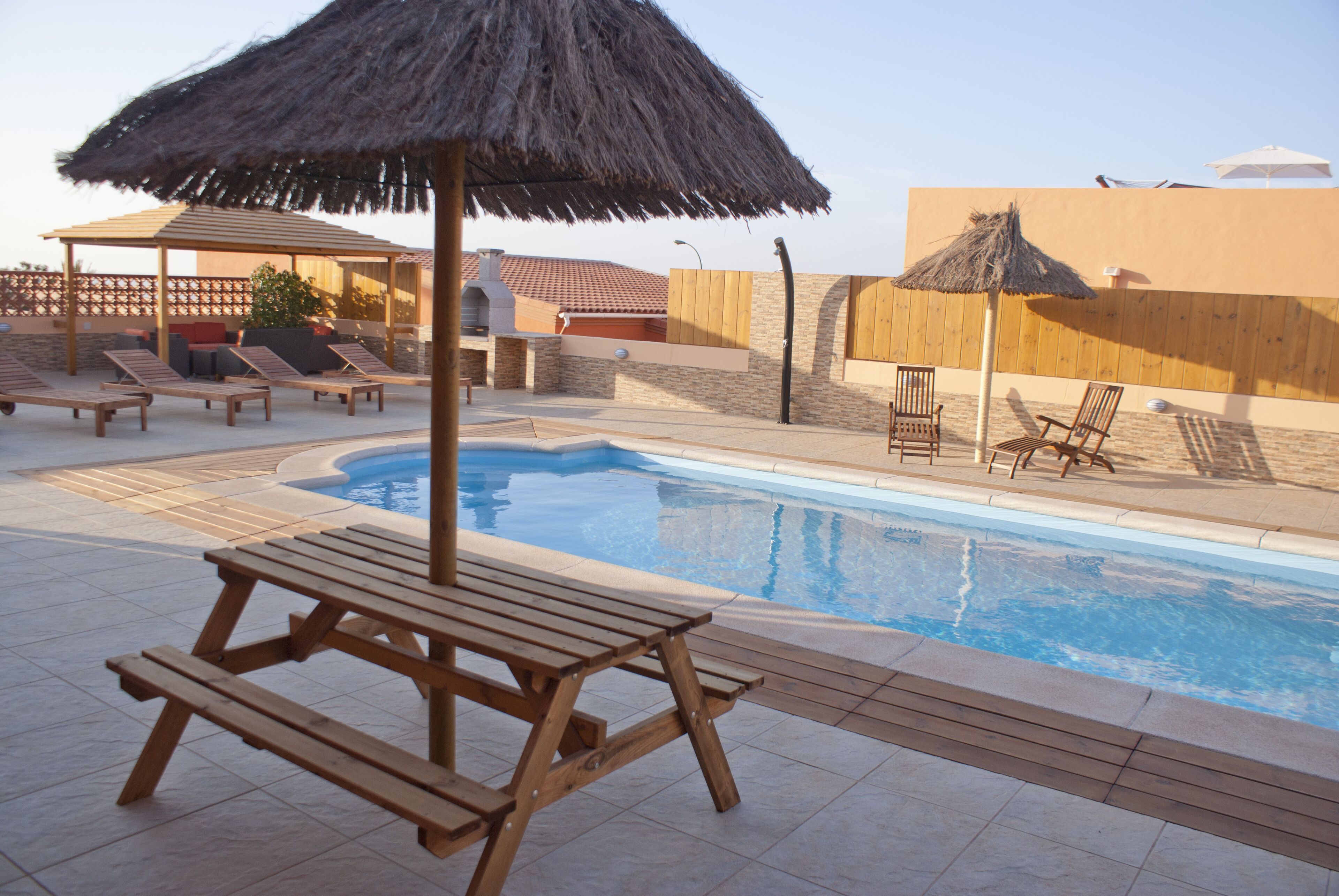 A pool area with wooden lounge chairs, a thatched umbrella, and a wooden table.