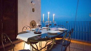 Una mesa de comedor dispuesta con velas, copas de vino y platos en un balcón con vista a un cielo azul.