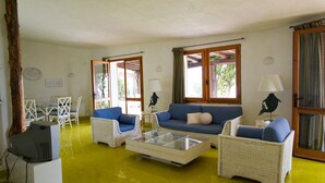 Uma sala de estar com móveis de vime, uma mesa de centro de vidro e um sofá com almofadas azuis. Há duas janelas com cortinas, uma porta e uma pequena mesa com abajur.