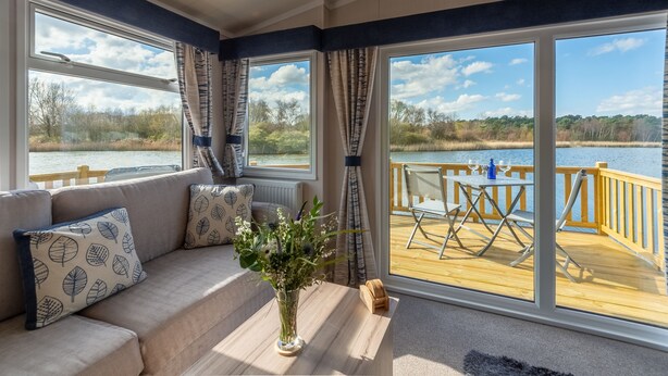 Ein gemütliches Zimmer mit einer Couch, einem Tisch mit Blumenvase und Blick auf eine Holzterrasse und Wasser.