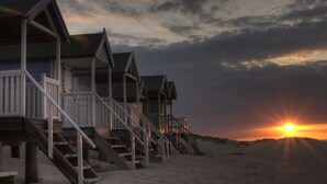Eine Reihe Strandhäuser mit Holzbalkonen und Treppen, die zum Strand führen, vor einem Sonnenuntergangshimmel.