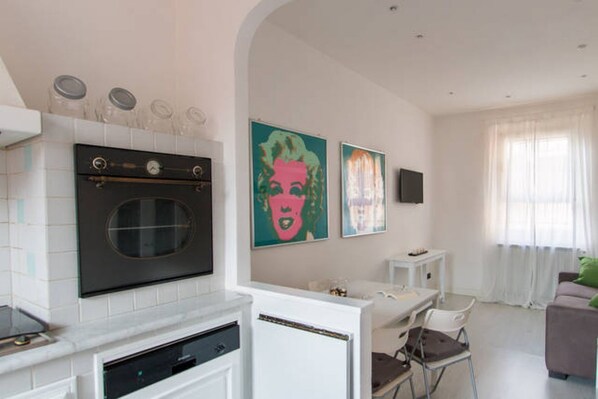 A modern kitchen with a built-in oven and dishwasher, a dining area with a white table and chairs, and a living room with a sofa and a TV.