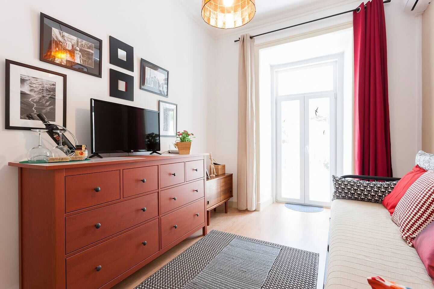 A modern bedroom with a TV, wooden dresser, and a large window with red curtains.