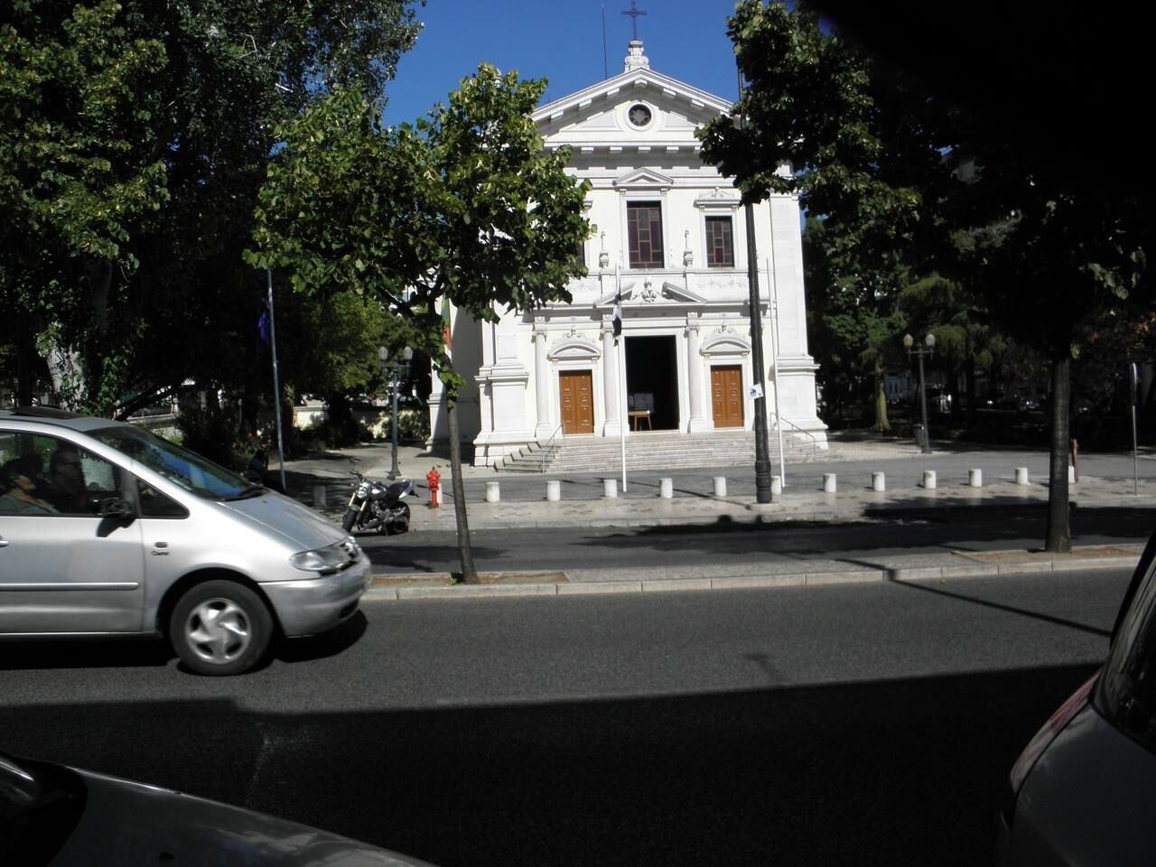 Un bâtiment blanc, doté d’une entrée centrale et surmonté d’une croix, entouré d’arbres et bordé d’une rue où circulent des voitures.