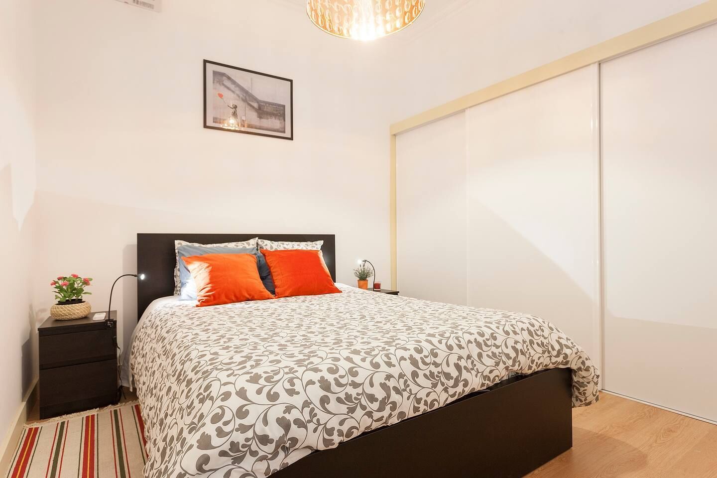Ein Schlafzimmer mit einem Bett, einem Nachttisch, einer Lampe, einer Topfpflanze und einem Bild an der Wand.