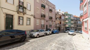 Eine enge Kopfsteinpflasterstraße, gesäumt von mehrstöckigen Gebäuden, parkenden Autos und mit Pflanzen bepflanzten Balkonen.