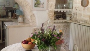 A rustic kitchen with stone walls, a wooden table with a lace tablecloth, a bowl of fruit, a vase with flowers, and a book.