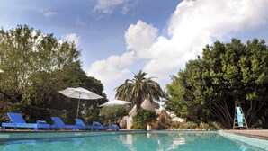 A swimming pool with blue lounge chairs and umbrellas, surrounded by trees and a stone structure.