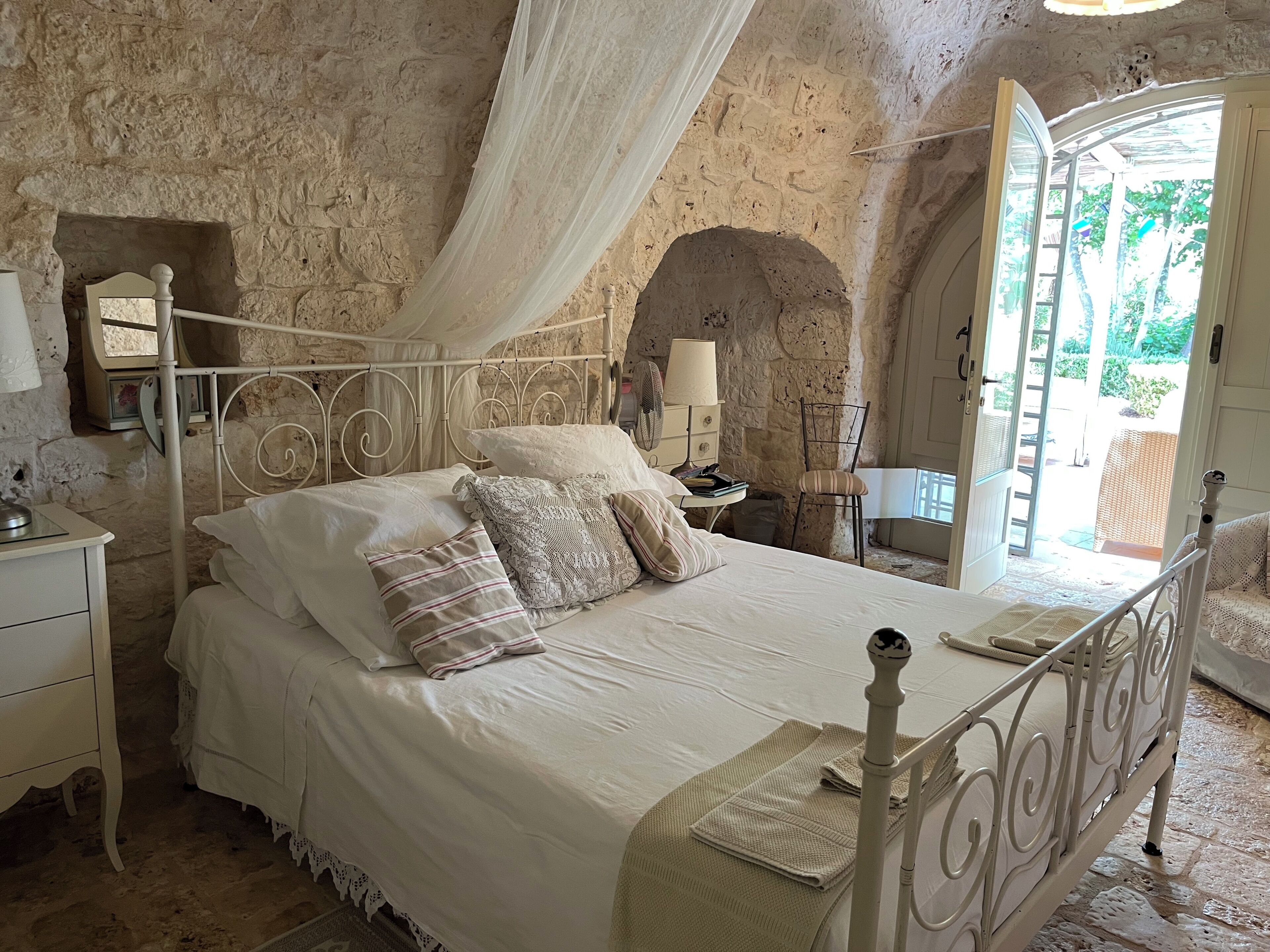 Ein Schlafzimmer mit Stehwänden, einem Bett mit weißer Bettwäsche, einem Nachttisch, einem Stuhl und einem Fenster mit Blick auf Grünflächen.