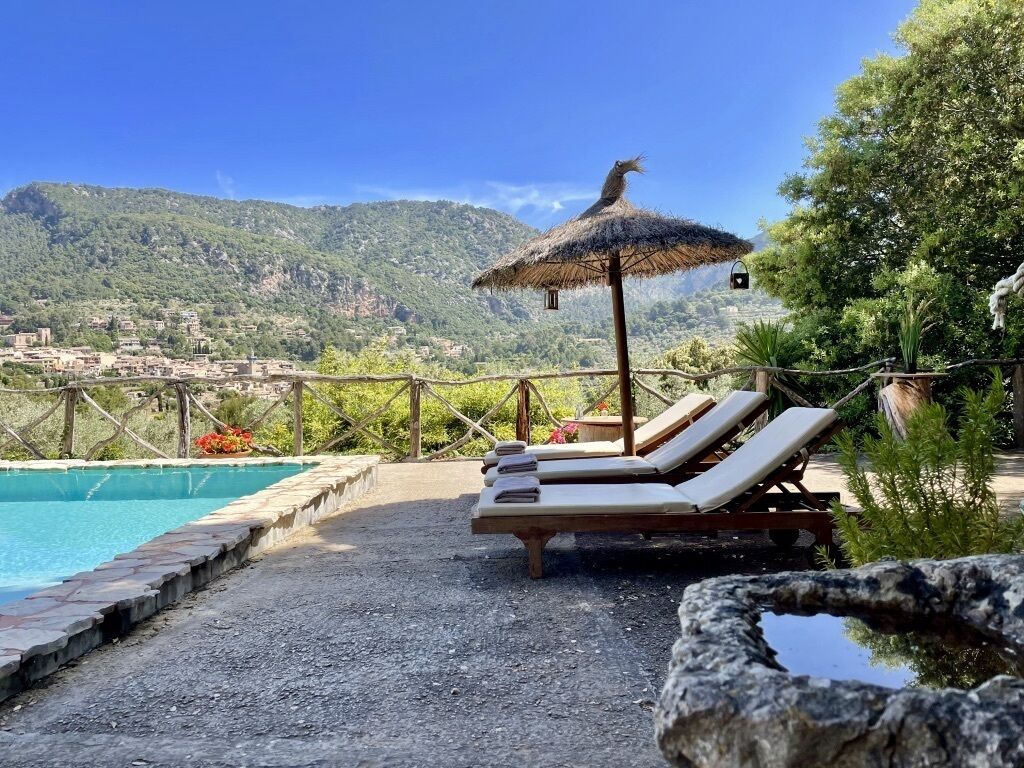 Zona de piscina con sillones de descanso y una sombrilla de paja, con vistas a un paisaje montañoso.