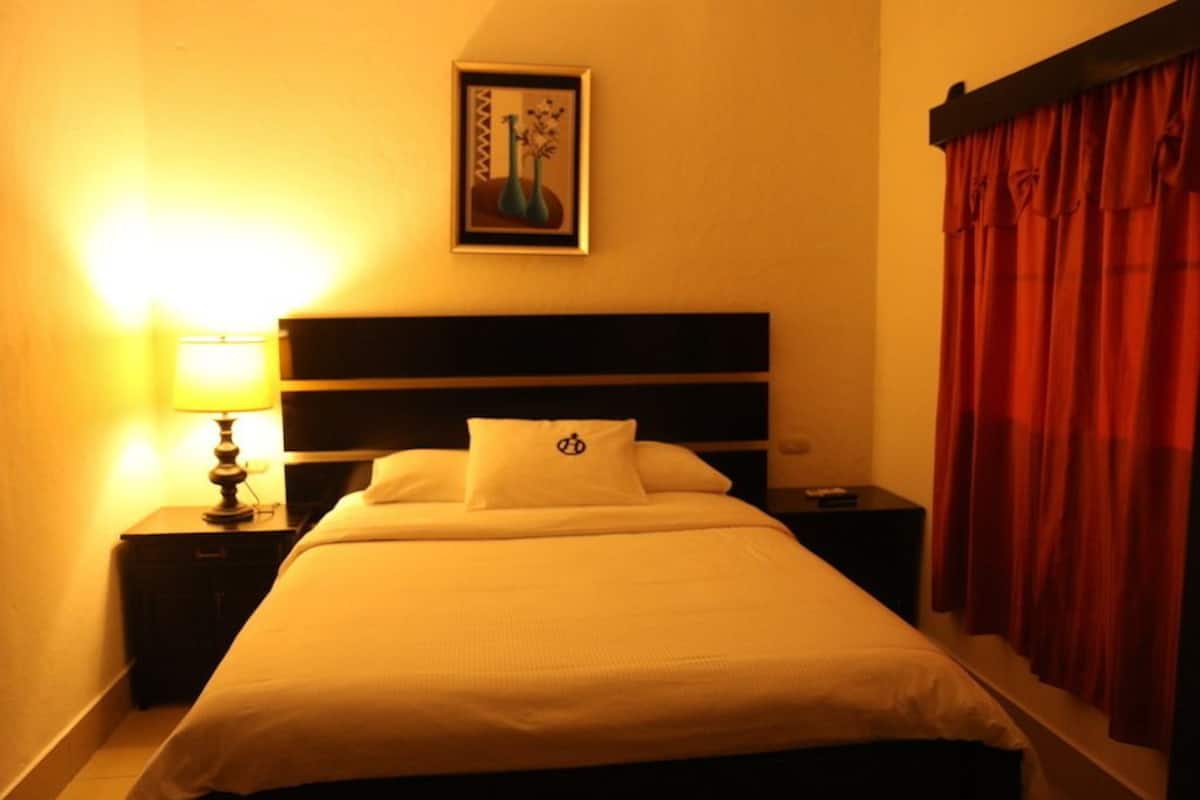 Een hotelkamer met een bed, een nachtkastje met een lamp, een ingelijst beeld aan de muur en rode gordijnen.