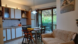 Uma sala de estar aconchegante com um sofá, uma mesa de jantar com cadeiras, uma cozinha com armários e uma janela grande com vista para o verde.