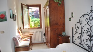 Quarto com cama, guarda-roupa de madeira, janela com vista para o verde, cadeira e radiador.