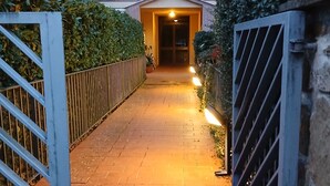 Uma passagem pavimentada leva a um acesso de edifício, com um portão à esquerda e uma área murada à direita.