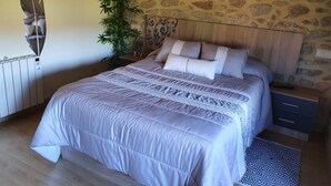 A bedroom with a stone wall, a bed with a patterned bedspread, a potted plant, and a wall-mounted picture of an orchid.