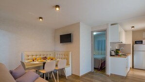 Un salon moderne avec un canapé beige, un coin repas avec une table et des chaises blanches, et une cuisine avec des meubles et des appareils électroménagers blancs.