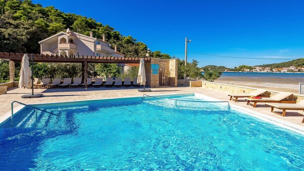 Une piscine dotée de chaises longues et d’une pergola, offrant une vue sur une plage et des collines.